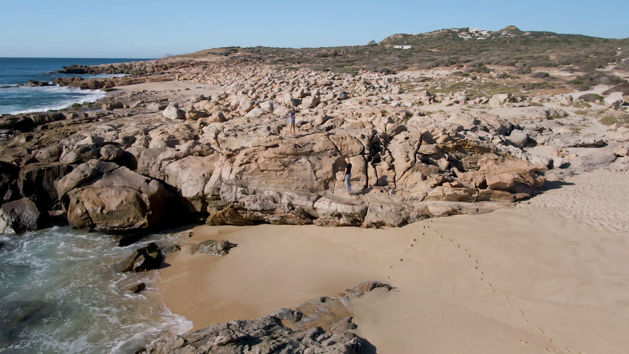 Aerial frame from San Jose Del Cabo, Mexico with a small wave reaching the sand, darker rocks near the water, broad tan boulders in the center, and tiny people visible among the rocks.