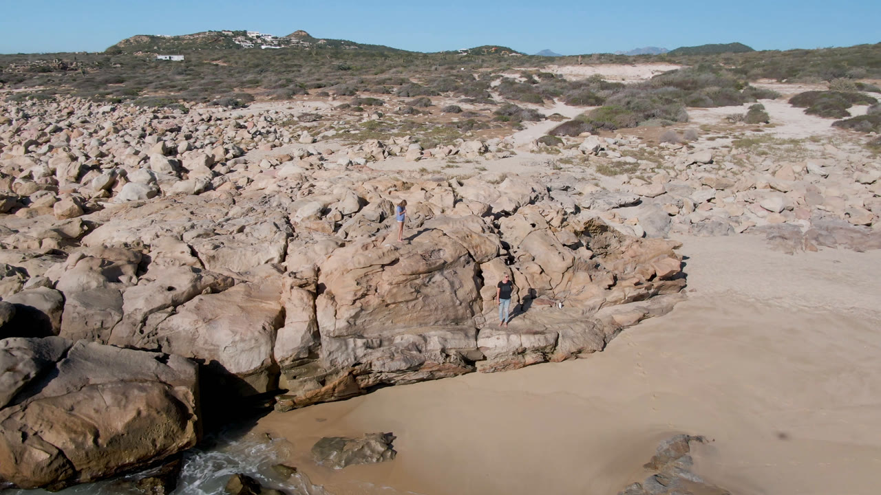 Aerial view of rocky beach terrain with sand, dunes, and distant hills.