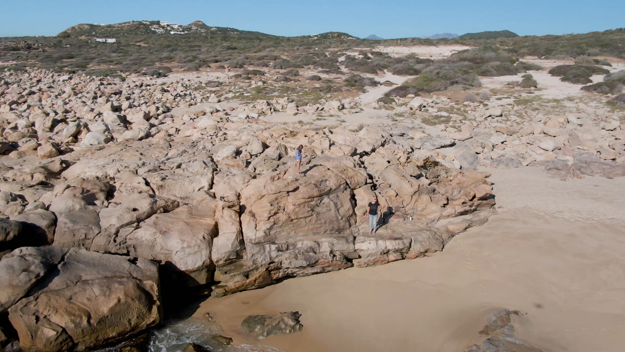 Aerial view over Laguna Beach rock formations in San Jose Del Cabo, Mexico, with clustered boulders, open sand, desert scrub, and tiny visitors standing among the rocks.