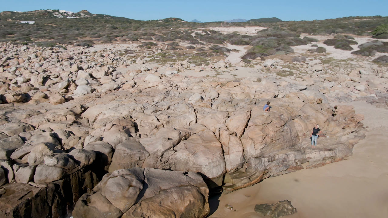 Aerial frame from San Jose Del Cabo, Mexico showing tan rock formations beside sand and scrub, with small distant people standing on the rocks under a clear blue sky.