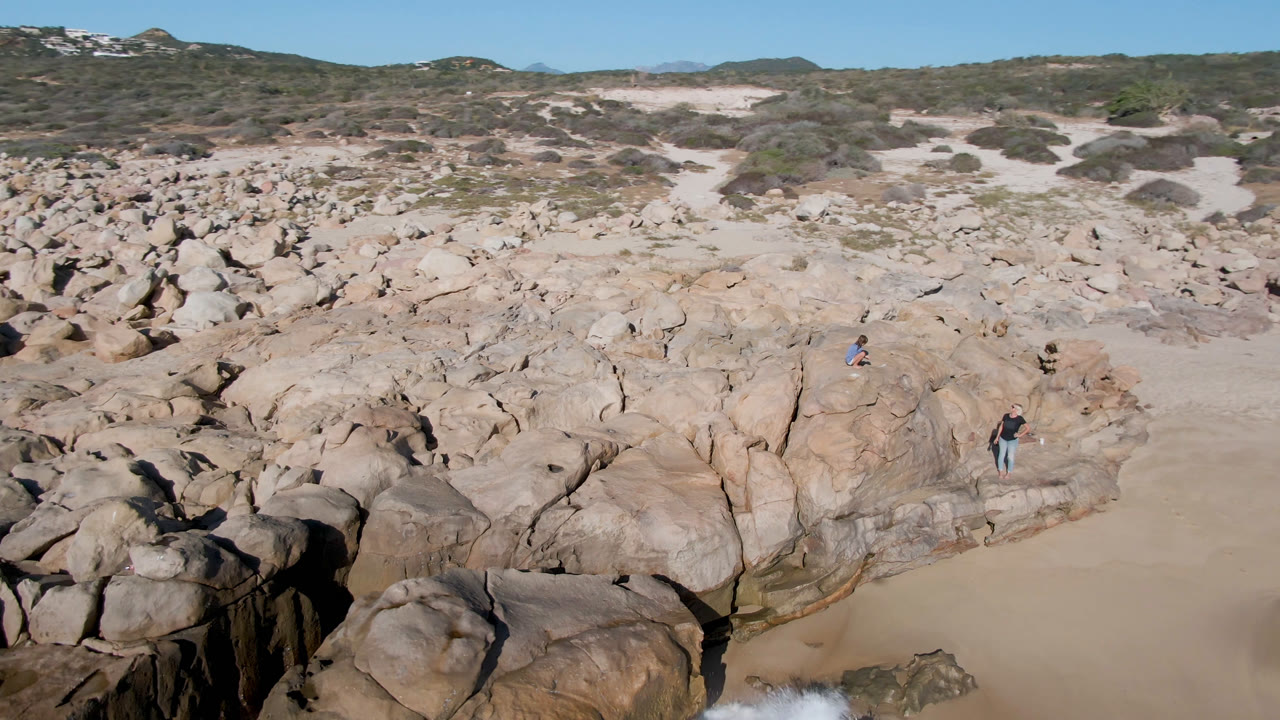 Aerial view in San Jose Del Cabo, Mexico with a broad rock plateau in the foreground, open sand behind it, scrub-covered dunes on the horizon, and distant people on the rocks.