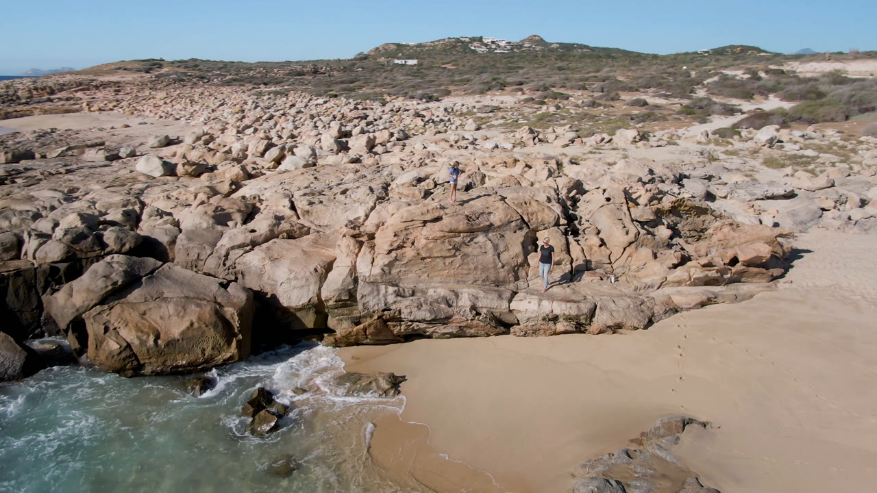 Aerial view of rocky beach with surf and small people on boulders.