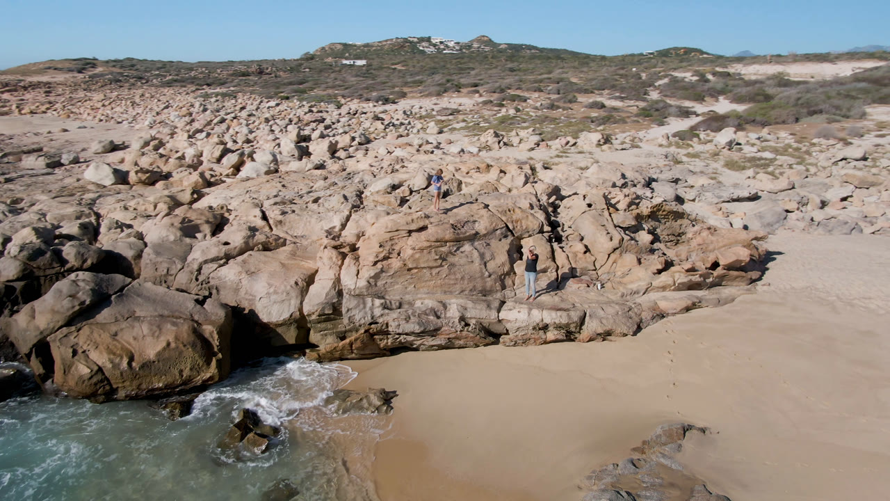 A second aerial frame of the San Jose Del Cabo coast shows the same boulder field and sandy cove, with whitewater shifting around the lower-left rocks and small people still visible on the formation.