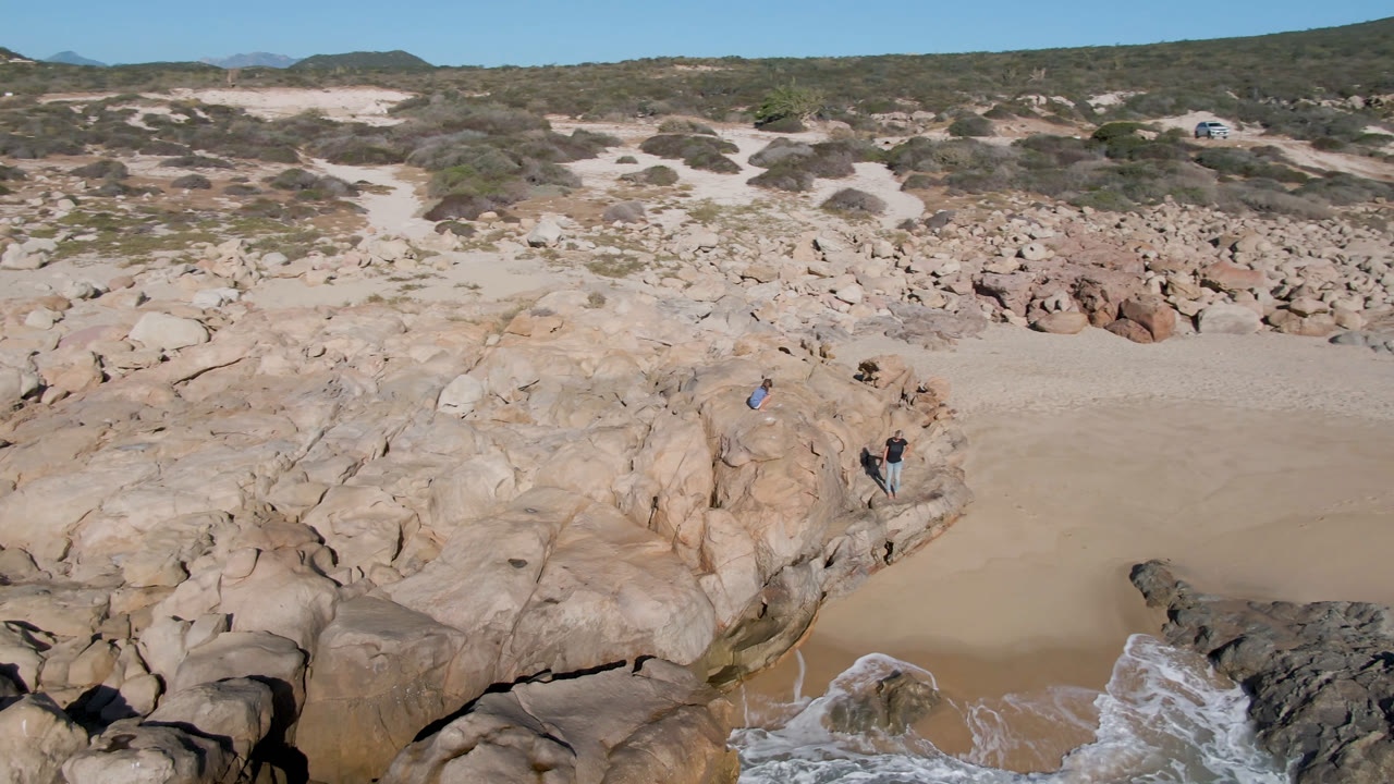 Aerial view of rocky beach with sand, surf, dunes, and small people on the rocks.