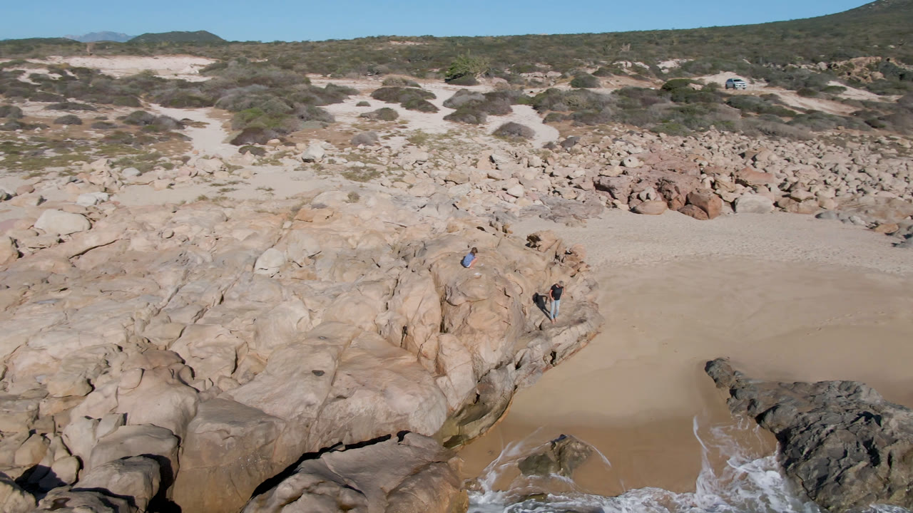 A second aerial view over San Jose Del Cabo, Mexico frames the rounded boulders more centrally, with a sandy beach behind them, surf below, and small people still visible on the rock face.