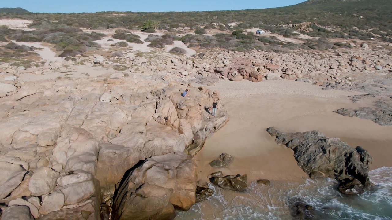Aerial frame from San Jose Del Cabo, Mexico with pale rocks in the foreground, an open sand pocket in the center, green scrub beyond, and a thin line of surf reaching the rocks with no visible people.