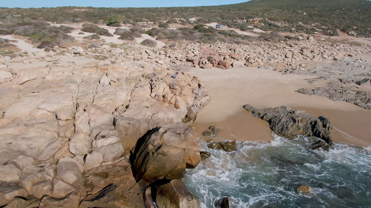 Elevated view of a rocky San Jose del Cabo beach cove with tan boulders, pale sand, turquoise surf, scrub hills, and small people visible near the rocks.