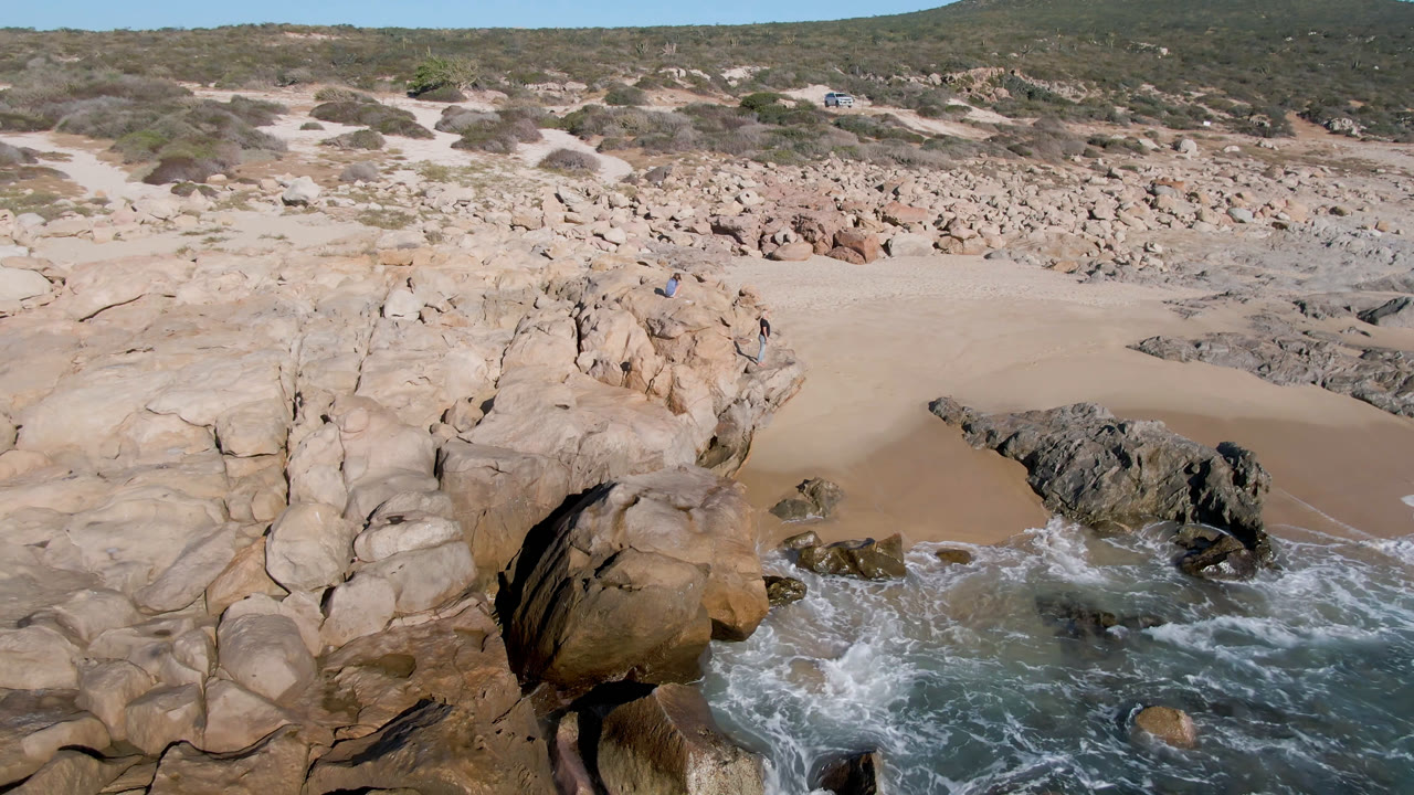 Aerial-looking frame over San Jose del Cabo rocks with white water curling around dark boulders, sandy beach behind, and tiny people visible near the upper rocks.