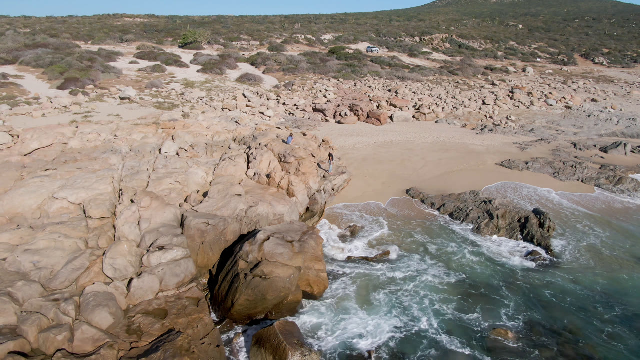 Aerial view of tan coastal rocks, surf, sand, and scrubland.