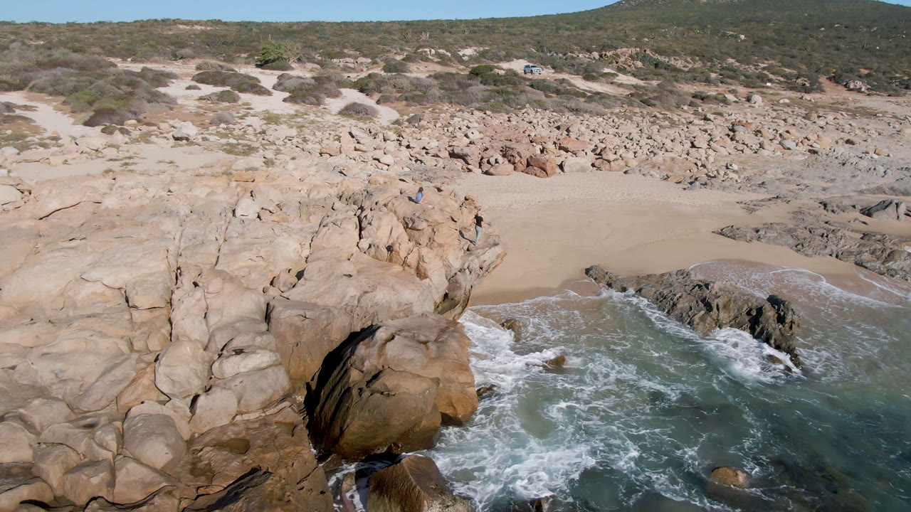 At 0:01, Laguna Beach Los Cabos appears from above with white water curling around foreground rocks, open sand beyond, low desert scrub, and no visible people.