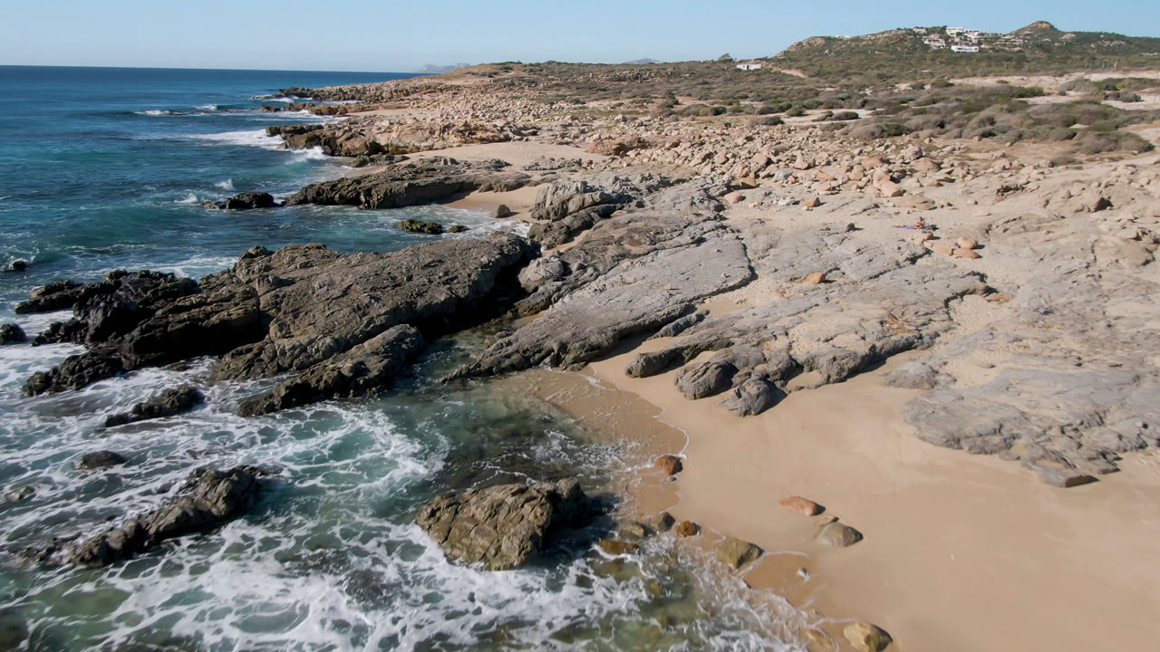 Laguna Beach Los Cabos shoreline seen from above, with foamy water pushing into a narrow sand pocket between dark foreground rocks and pale stone slabs.