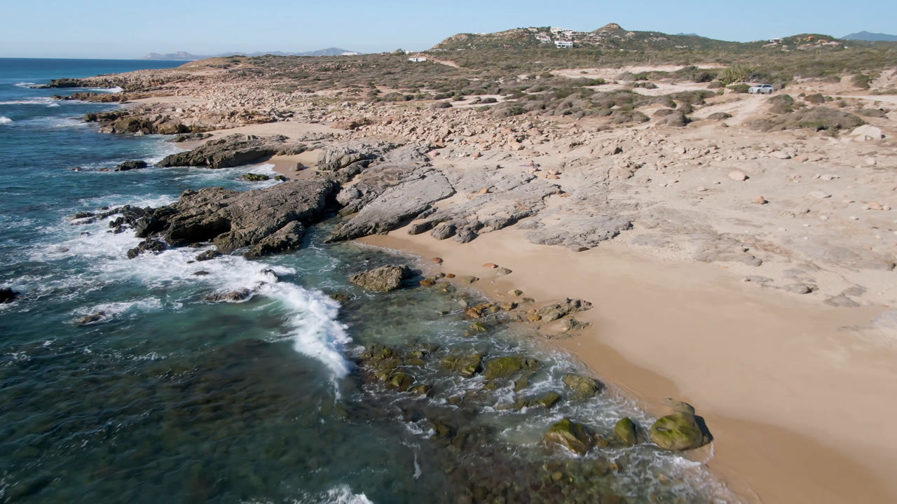 Aerial view of Laguna Beach Los Cabos with blue surf entering from the left, a sandy beach on the right, rocky shoreline ahead, scrub hills in the background, and no visible people.