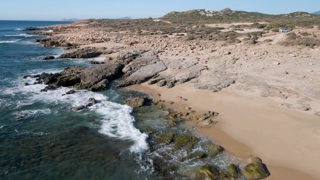 Aerial frame over Laguna Beach Los Cabos where a white wave curls across dark rocks beside the tan beach, with low coastal hills and no visible people.