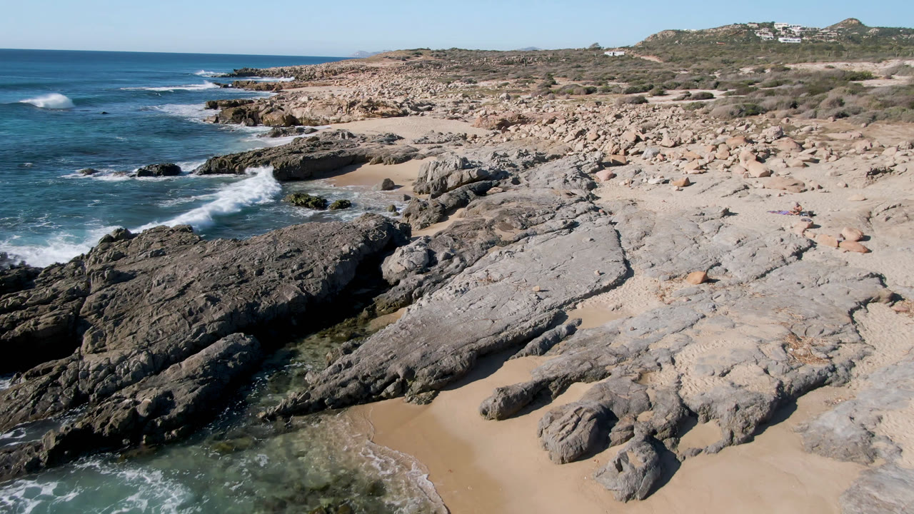 Elevated view of Laguna Beach Los Cabos showing dark foreground rocks, pale stone shelves, blue surf, sandy pockets, distant hillside buildings, and tiny beachgoers.