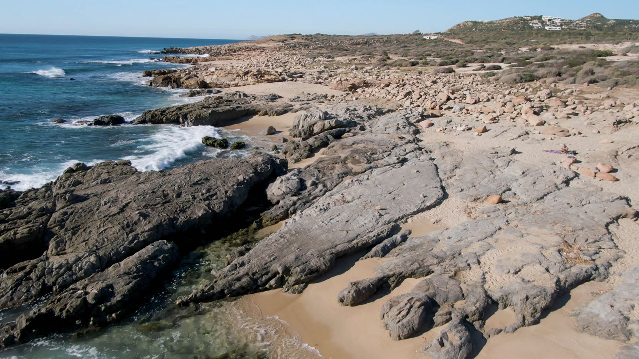 Laguna Beach Los Cabos shoreline with a larger white wave breaking near the left rocks, broad pale stone in the center, sand below, and distant hillside structures.