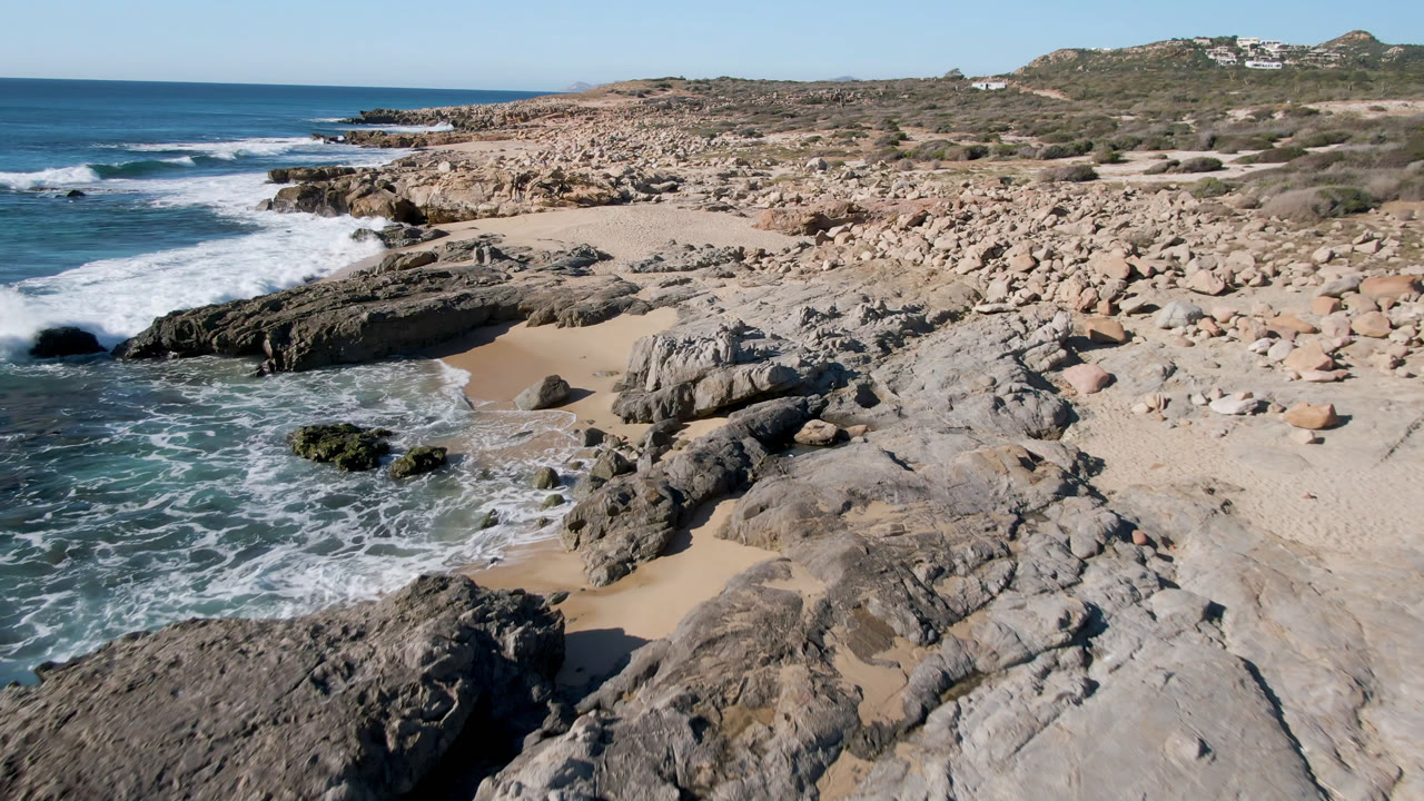 Elevated view of Laguna Beach Los Cabos with dark rock ledges, tan sand, blue surf on the left, boulder-covered shore, distant hillside buildings, and no visible people.