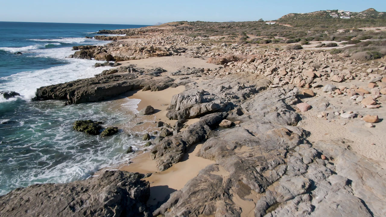 Laguna Beach Los Cabos shoreline seen from above as foam spreads into a sandy pocket between gray rock shelves, with boulders and low hillside behind and no visible people.