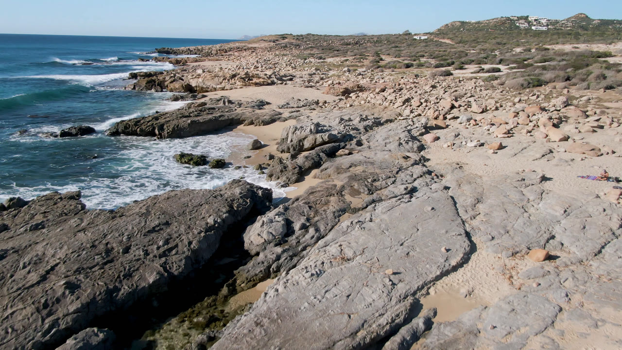 Elevated view of Laguna Beach Los Cabos with blue ocean on the left, gray rock shelves in the foreground, pale sand beyond, scrubby hillside in back, and no visible people.