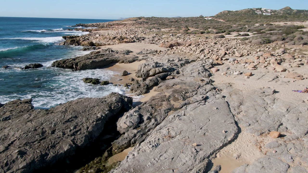 Laguna Beach Los Cabos shoreline seen from above, with a wave line approaching dark rocks, a small sandy pocket at center, dry scrub inland, and no visible people.