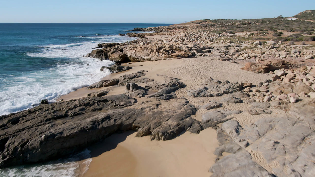 Elevated view of Laguna Beach Los Cabos with blue surf on the left, pale sand below, dark foreground rocks, scattered tan boulders, and no visible people.
