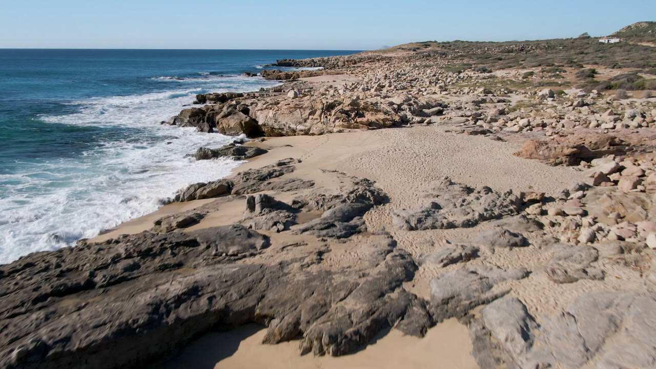 Laguna Beach Los Cabos shoreline with white surf curling into a sandy cove, dark rocks in the foreground, a boulder field beyond the beach, and no visible people.