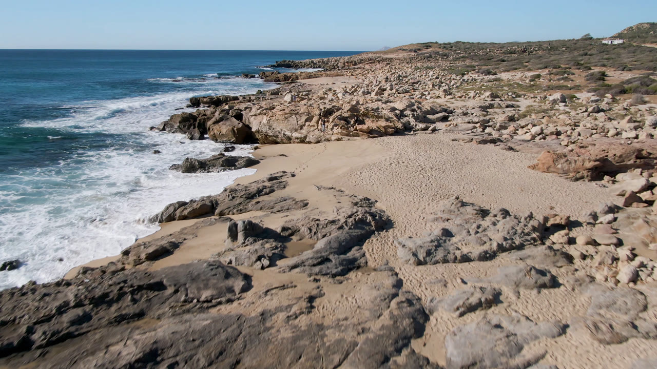 A wide aerial frame over Laguna Beach Los Cabos shows the sandy beach centered between foaming surf, dark foreground rock, tan boulders, low scrub, and no visible people.