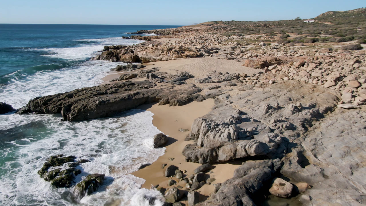 Laguna Beach Los Cabos shoreline with a larger band of whitewater breaking beside dark rocks, exposed sand in the cove, open sea in back, and no visible people.