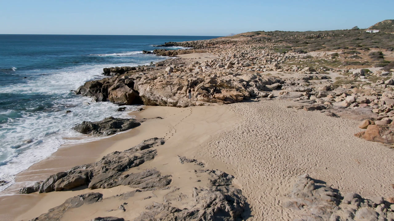 Elevated view of Laguna Beach in San Jose Del Cabo, Mexico, with a sandy cove, dark shoreline rocks, white surf at left, low coastal scrub, and no clearly visible foreground people.