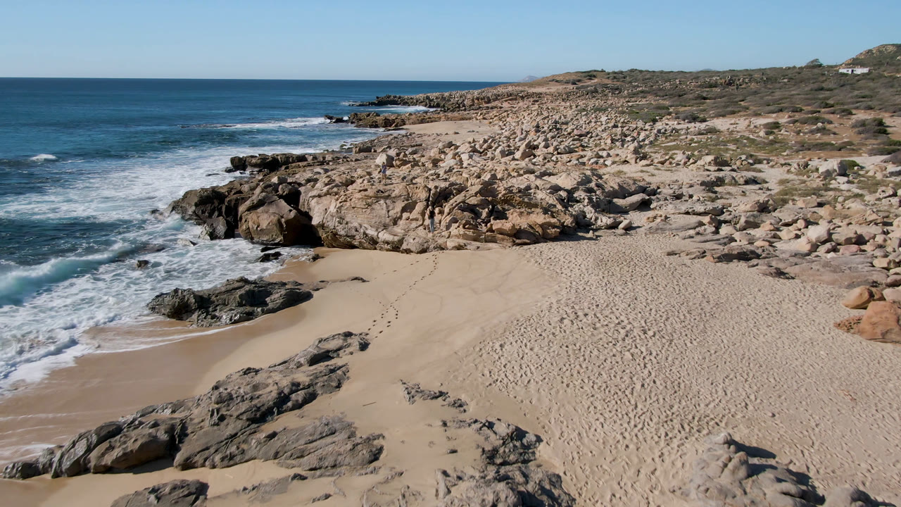 Aerial-style frame over Laguna Beach in San Jose Del Cabo, Mexico, showing footprints across pale sand, waves pushing around black rocks, tan boulders on the point, and a bright blue horizon.