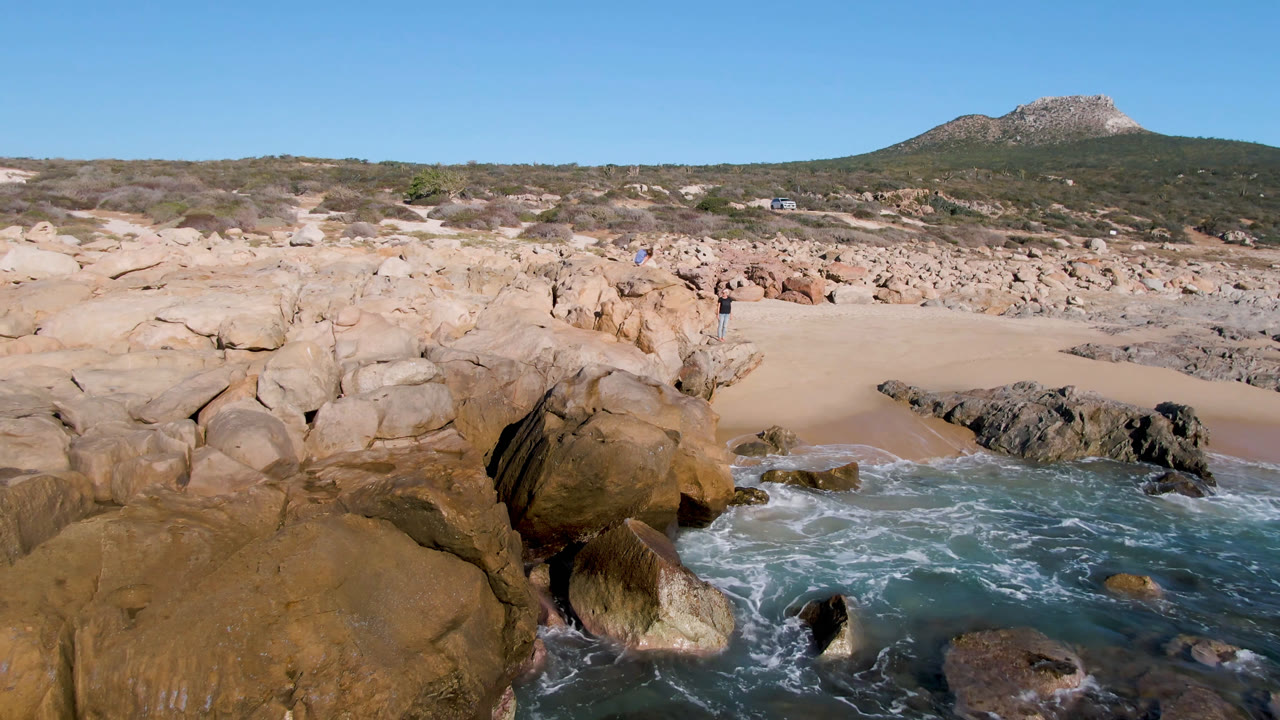 La Laguna Beach in Los Cabos with tan foreground boulders, turquoise water entering a sandy cove, scrub-covered hillside in back, and no visible people.