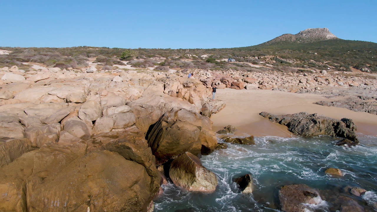 La Laguna Beach shoreline with foamy water curling around dark midground rocks, pale sand behind them, and a low green hillside under clear sky with no visible people.