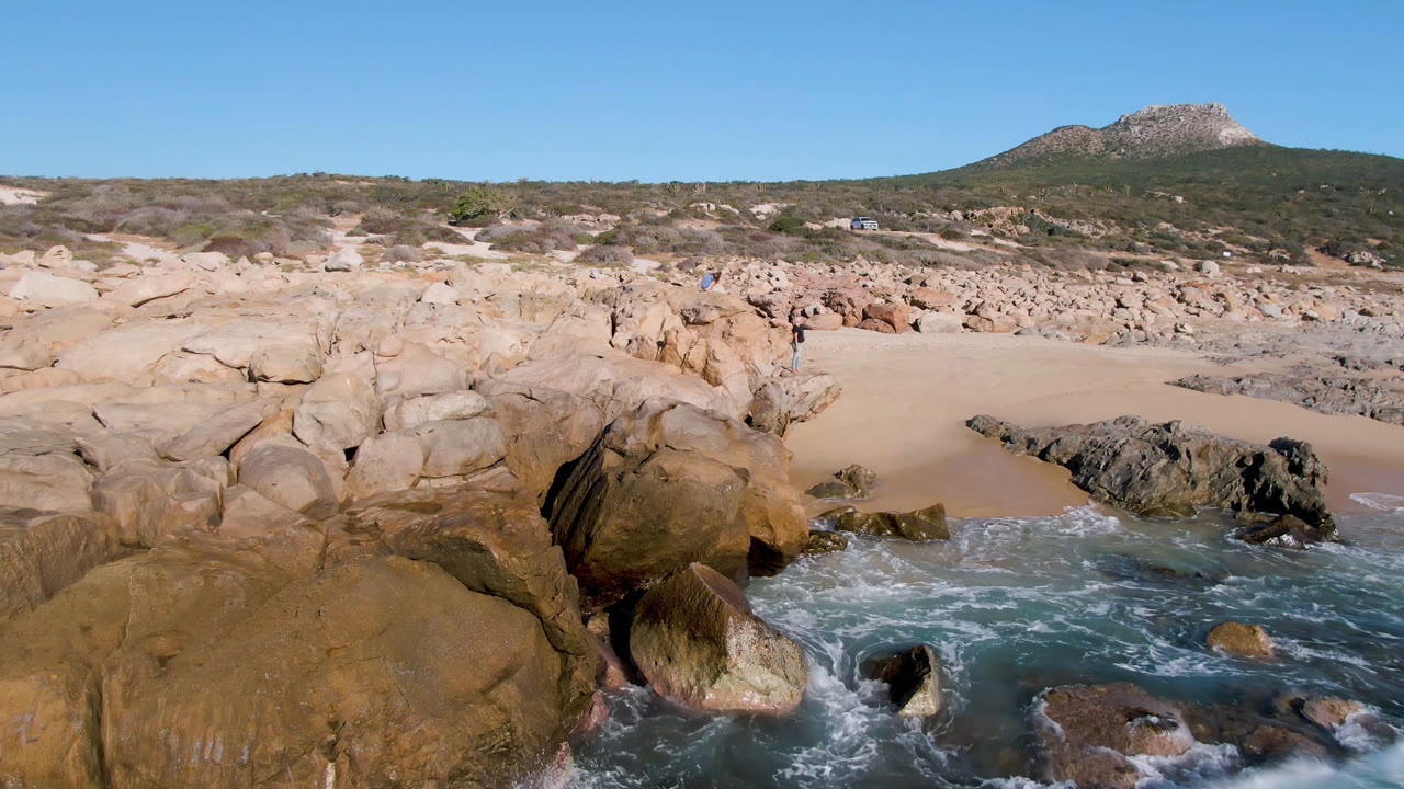 Rocky coastal inlet with small waves, sand, scrub hills, and clear sky.