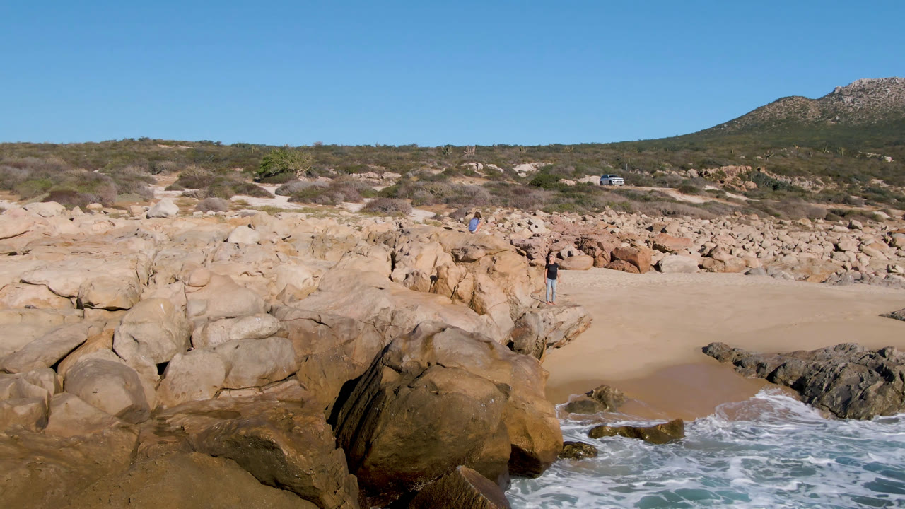 Rocky sandy beach with surf, scrub hills, and small distant people.