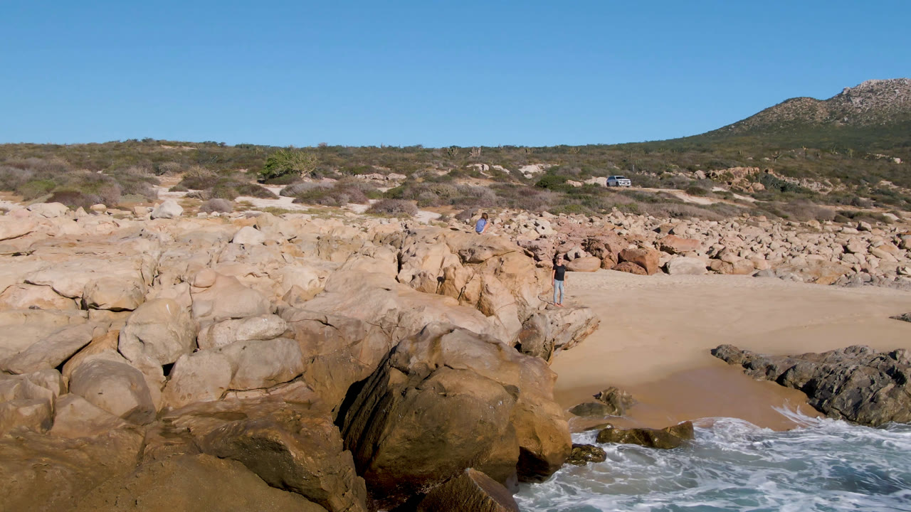 Laguna Beach Los Cabos shoreline with a broader wash of whitewater at the bottom edge, tan rock formations around the sand, scrubland, a hill, and small people visible in the distance.