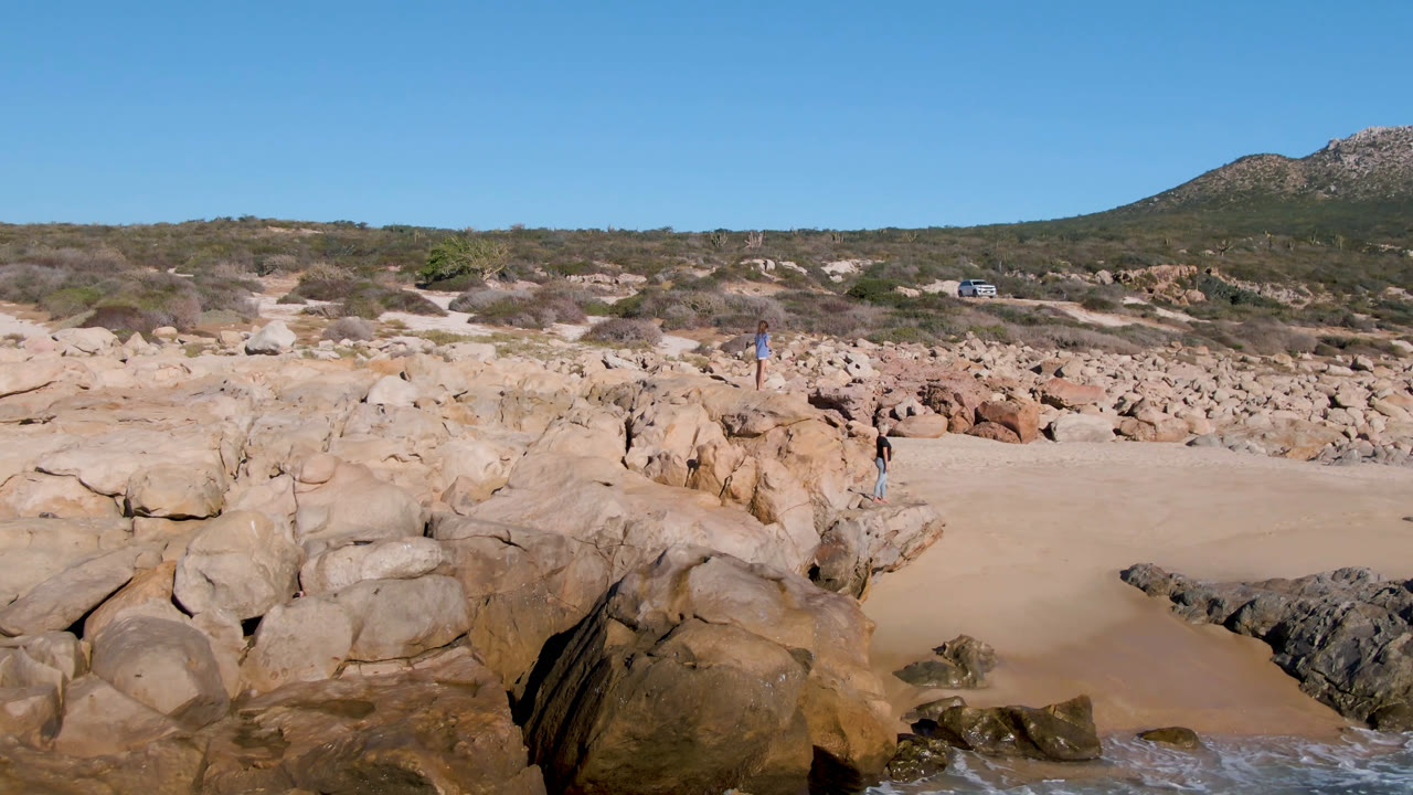 Opening frame at Laguna Beach Los Cabos shows tan foreground rocks, a sandy cove, small people on the rocks, scrub-covered dunes, a rocky hill, and blue sky.