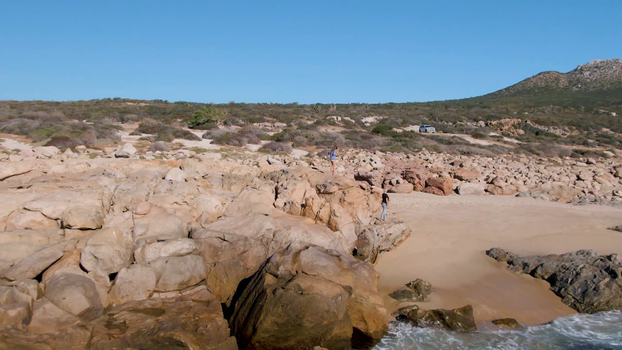 At 0:01, the Laguna Beach Los Cabos view holds on the sandy cove between large rocks, with distant people still visible on the rocks and surf breaking at the lower edge.
