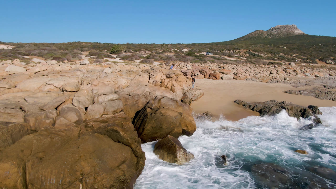 Wide view of Laguna Beach Los Cabos with foamy water around foreground boulders, a sandy cove, scrub-covered hills, and small people visible near the rocks.
