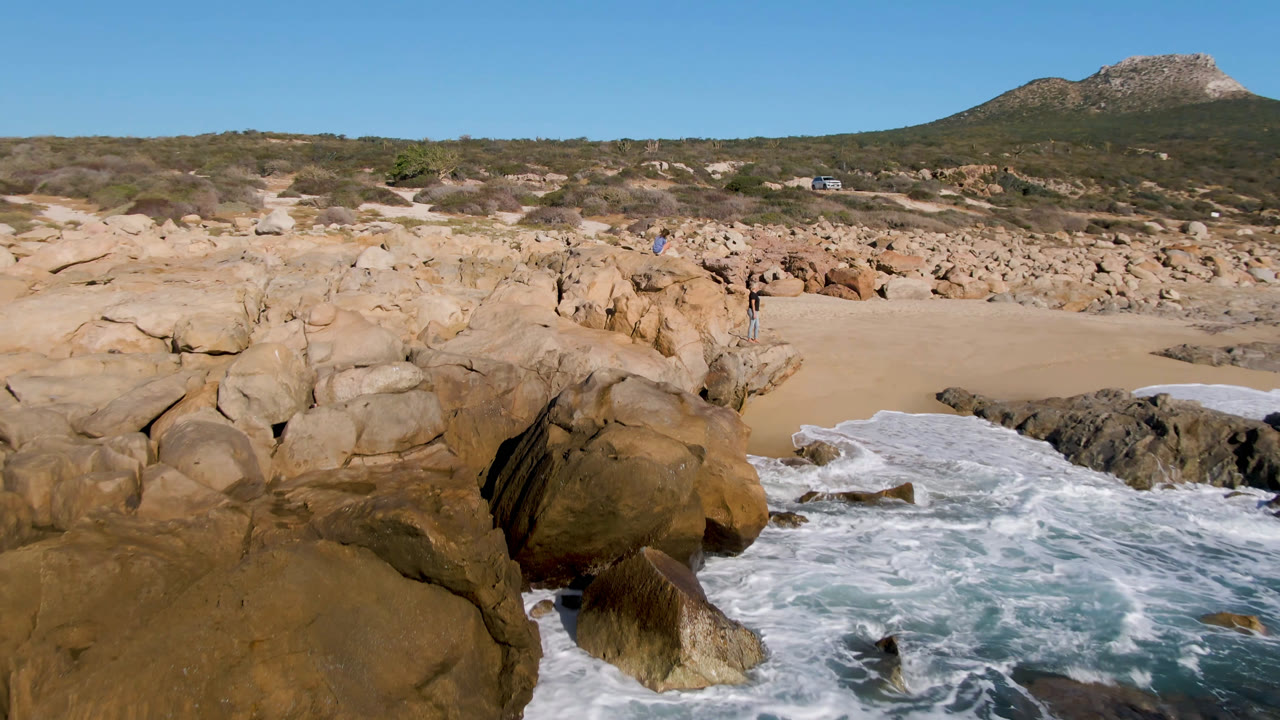 A wide Laguna Beach Los Cabos frame with surf curling around dark foreground rocks, tan boulders at center, beach sand behind them, and small people visible against the rocky midground.
