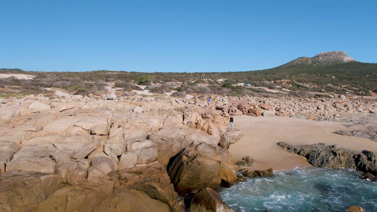 Rocky beach with blue-green water, sand, scrub hills, and clear sky.