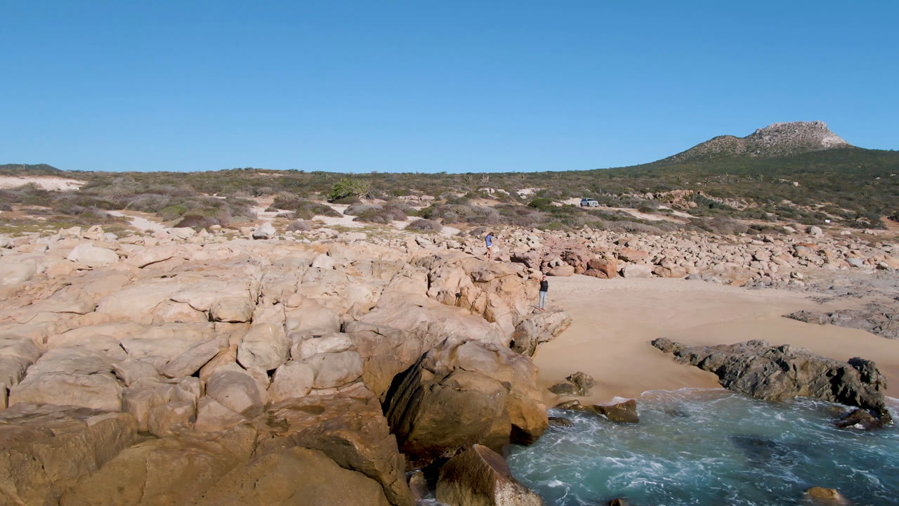 Laguna Beach Los Cabos shoreline seen from above, with white foam shifting around dark rocks in the foreground and a sandy pocket below low desert vegetation and a rocky hill.