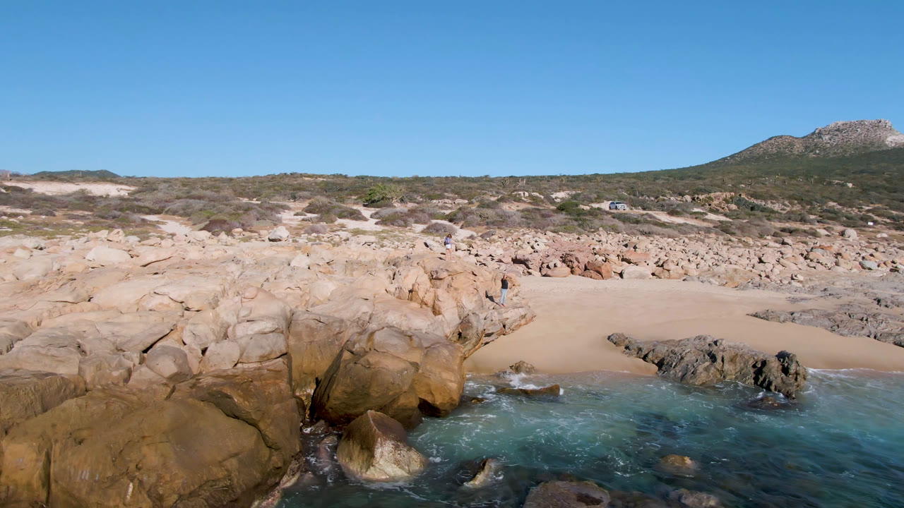 Laguna Beach Los Cabos rocky cove with tan boulders in the foreground, a small sandy beach beyond them, low hills in the distance, and no visible people.
