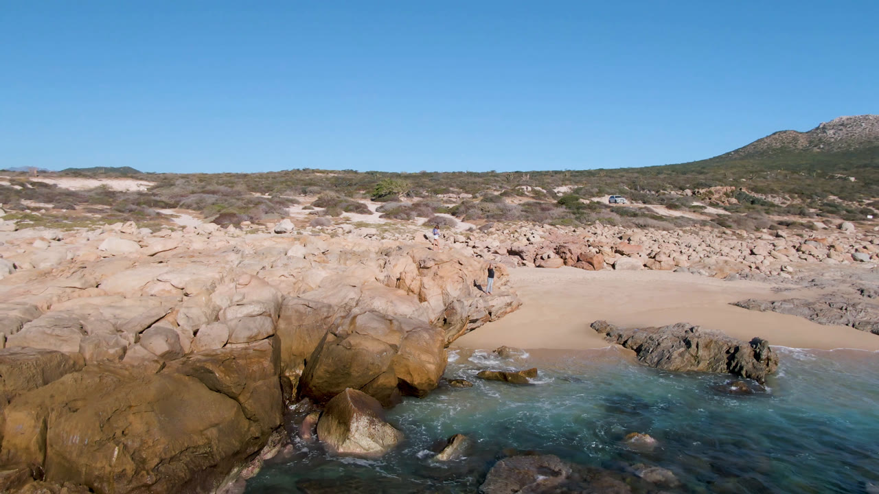 Laguna Beach Los Cabos shoreline with shallow blue-green water around foreground rocks, pale sand in the midground, scrub-covered dunes, distant hills, and no visible people.