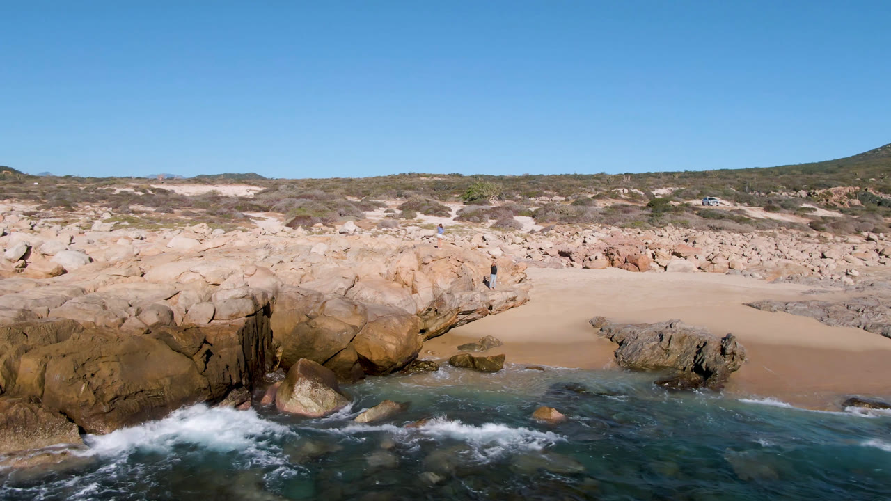 Laguna Beach Los Cabos shoreline with turquoise water, tan rocks, pale sand, low dunes, and a distant person standing on the cliff at the start of the clip.