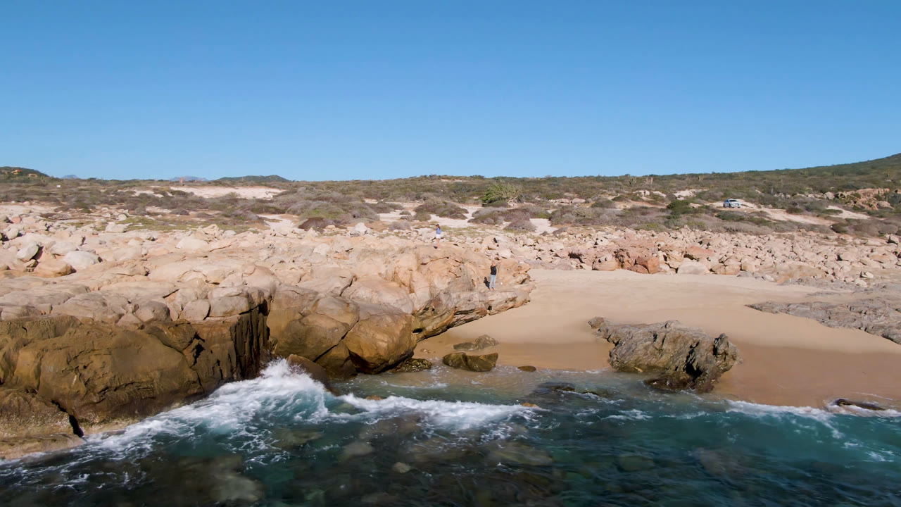 At 0:01, surf pushes harder into the rocks below the sandy Laguna Beach Los Cabos cove, with the distant person still visible on the cliff.