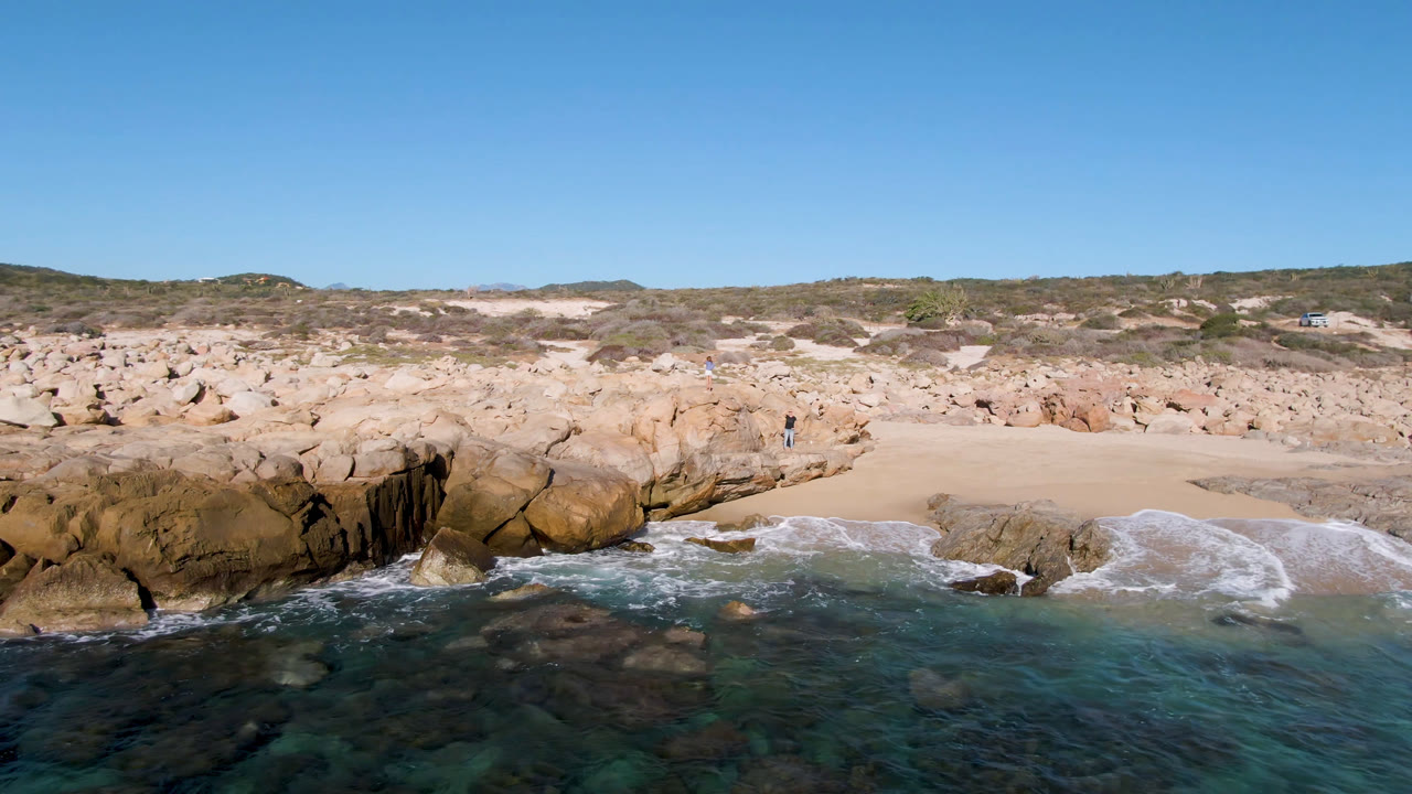 Wide view of Laguna Beach in Los Cabos with clear water in the foreground, tan rocks and sand across the shore, and a distant person standing on the rocky outcrop.