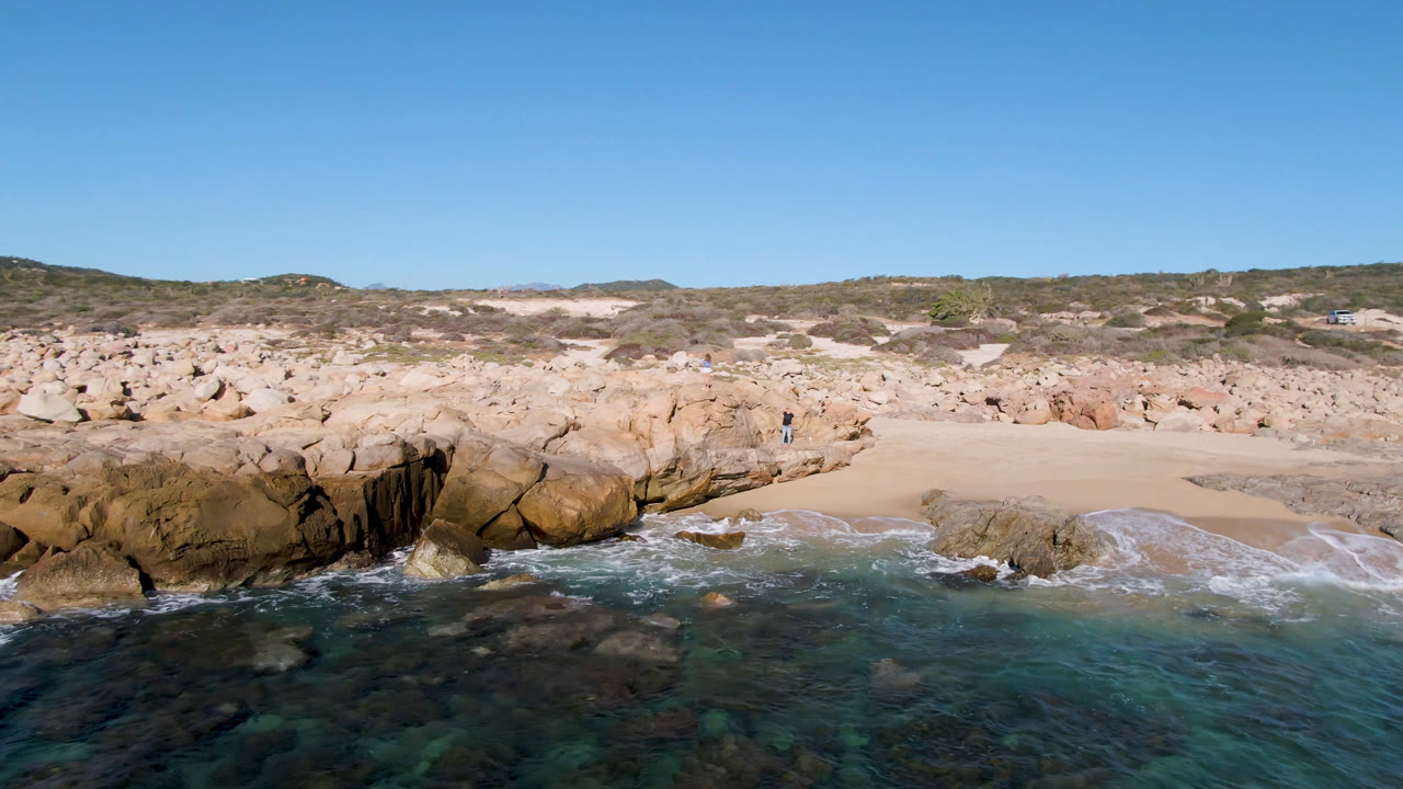 Ocean wash against boulders and sand with a distant person on the rocks