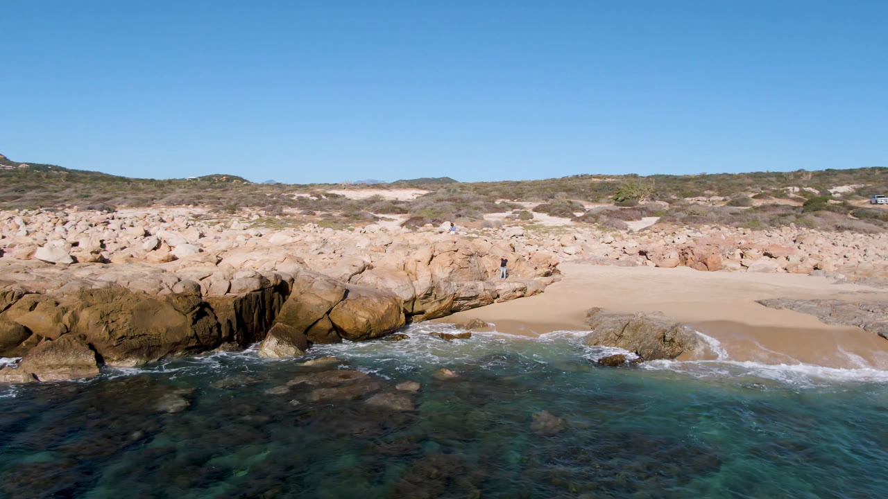 A wide aerial-looking frame of Laguna Beach shows shallow blue-green water, dark rocks below the surface, a tan boulder line, open sand, dunes, and a small person on the rocks.