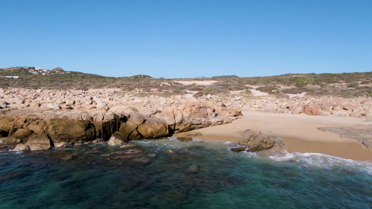 Opening view of Laguna Beach Los Cabos with clear water in the foreground, tan rocks, pale sand, low hills, and no visible people.