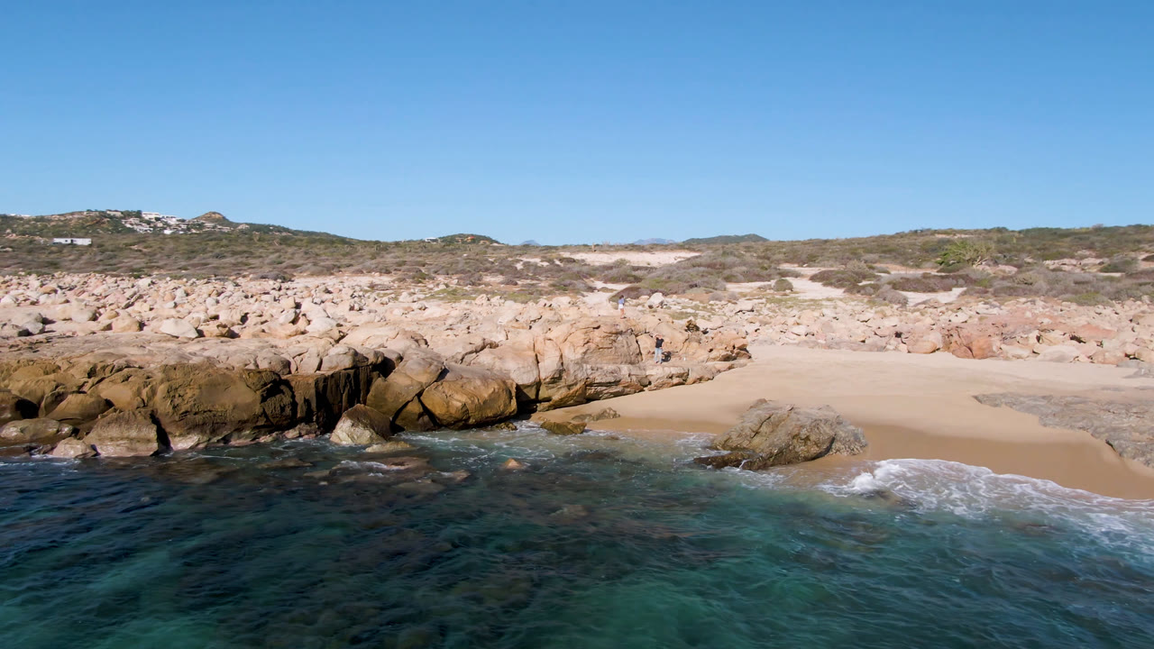 Laguna Beach Los Cabos shoreline with a thin wave line at the sand, large tan rocks across the center, blue-green water, and no visible people.