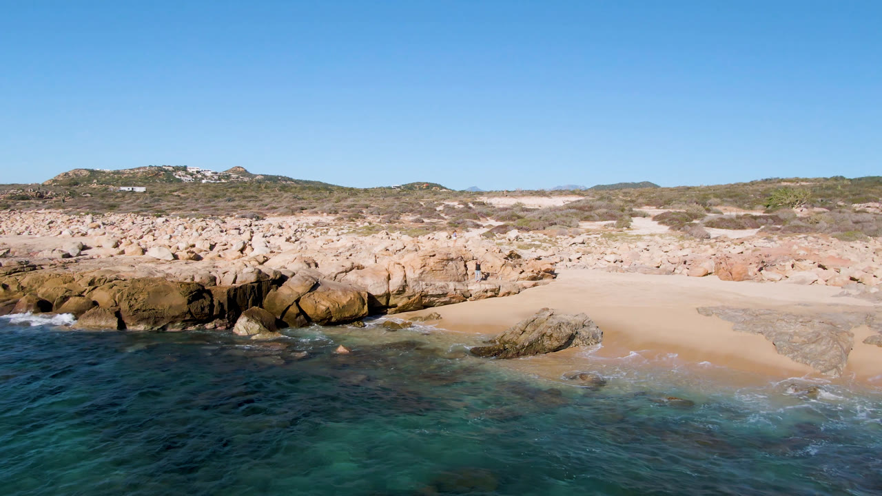 Wide opening frame of Laguna Beach Los Cabos showing blue ocean in the foreground, a pale sand pocket, tan rock ledges, scrubby hills, and no clearly visible people.